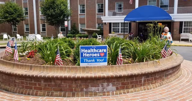 A circular brick planter filled with green plants and small American flags in front of a senior living facility building. A blue sign in the planter reads 'To Our Healthcare Heroes Thank You!'. There are white chairs and a blue awning at the building entrance. An elderly woman stands near the planter.