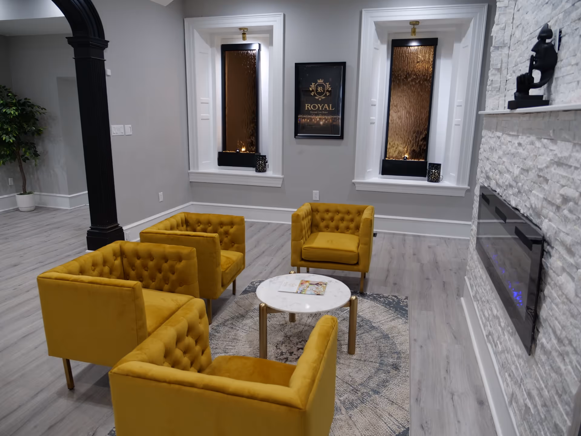 A modern lounge area with four mustard yellow tufted armchairs arranged around a small round white marble table on a patterned rug. The room has light gray walls, two tall windows with textured glass, a black framed 'Royal Personal Care Homes' sign between the windows, a white stone accent wall with a mounted electric fireplace, and a black decorative sculpture on a white mantel.