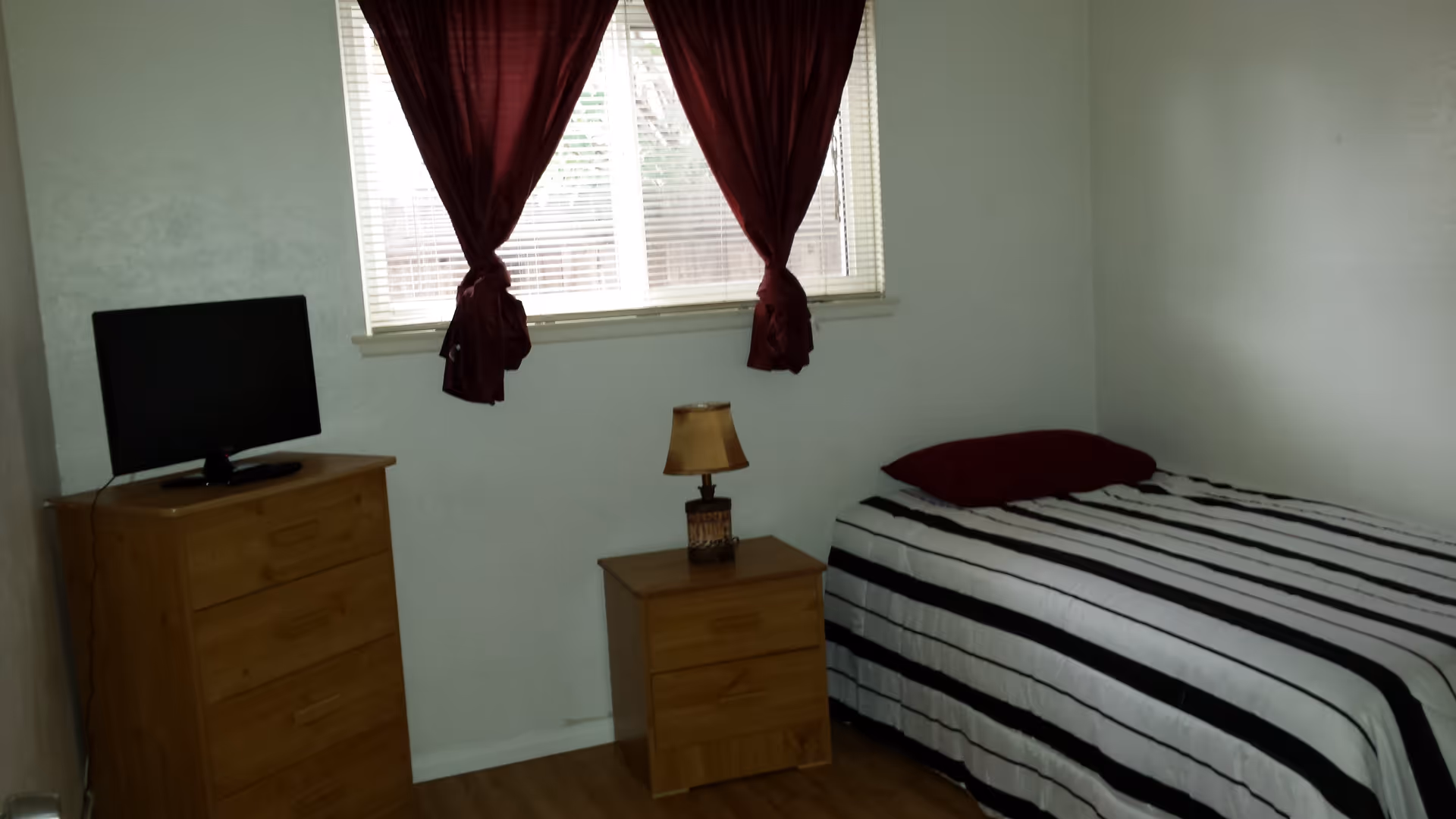 A small bedroom with a single bed covered in a black and white striped bedspread and a dark red pillow. Next to the bed is a wooden nightstand with a small lamp. Across from the bed is a wooden dresser with a flat-screen TV on top. A window with white blinds and dark red curtains is above the nightstand.