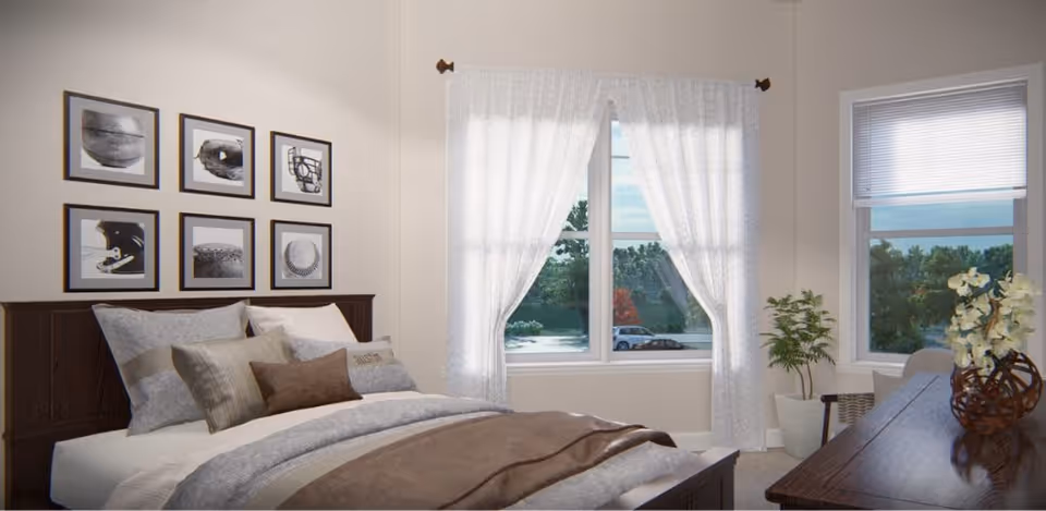 A cozy bedroom with a neatly made bed featuring multiple pillows and a brown blanket. Above the bed, there are six framed black and white pictures arranged in two rows. The room has two windows with white curtains and blinds, letting in natural light and showing a view of trees and a parked car outside. A wooden dresser with a decorative vase and flowers is positioned near one window, and a potted plant is placed on the floor beside it.
