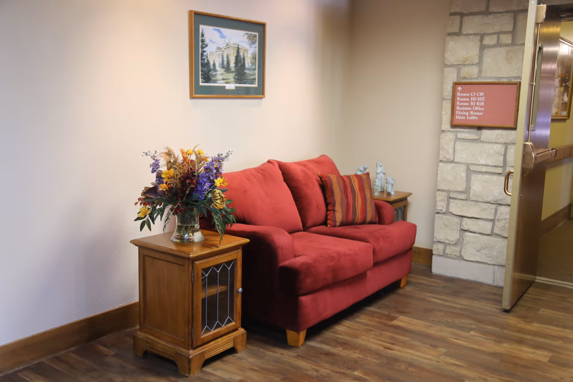 A cozy corner in a facility with a red upholstered loveseat adorned with a striped cushion. To the left of the loveseat is a wooden side table with a glass vase holding a colorful bouquet of flowers. On the right side, another wooden side table holds three small decorative figurines. Above the loveseat hangs a framed painting of a building surrounded by trees. A stone wall with a directional sign and an open door leading to a hallway are visible on the right side.
