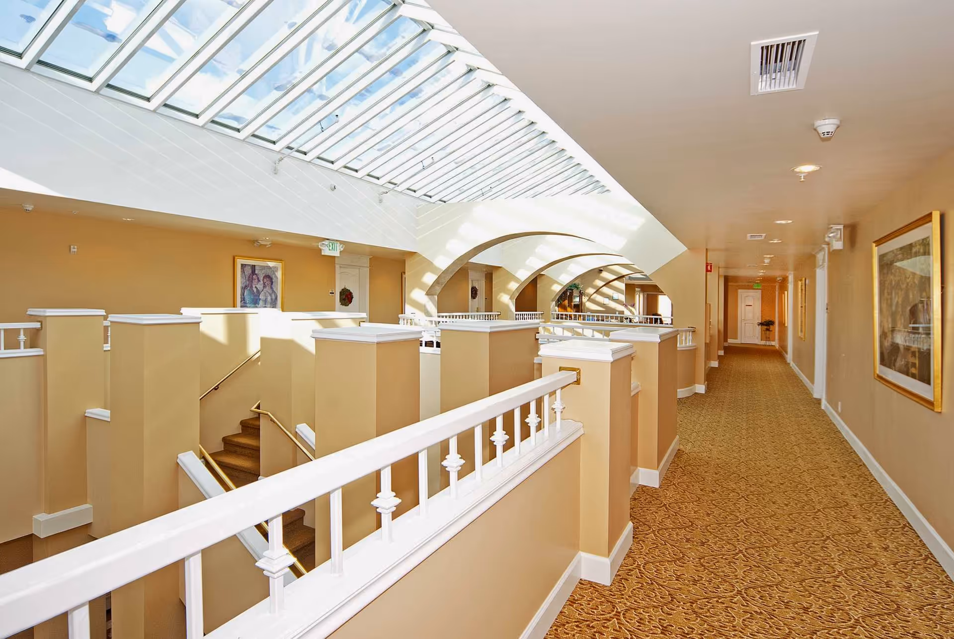 Bright, carpeted interior hallway with white railings, arched skylights, and framed artwork.