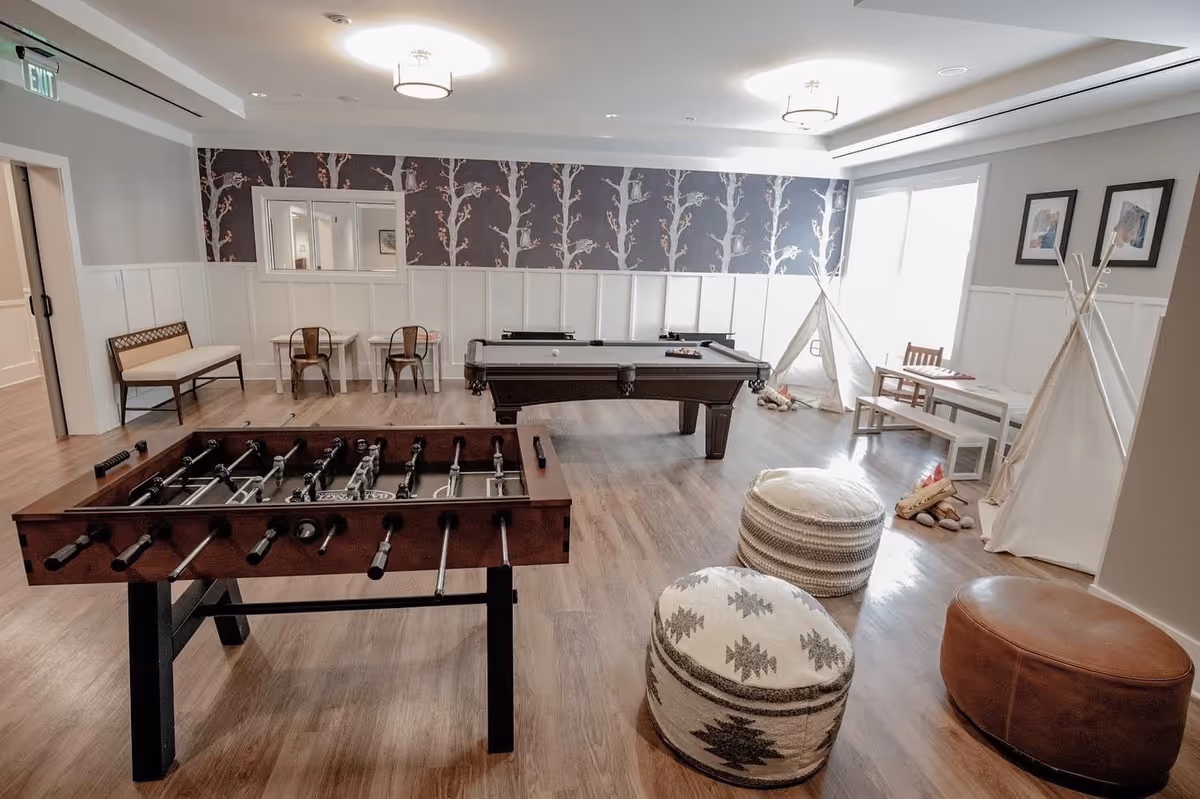 A spacious recreational room with a foosball table in the foreground and a pool table in the center. The room has wooden flooring, patterned wallpaper with tree designs, and white wainscoting on the walls. There are two small teepees with cushions and toys inside, a small table with chairs, and two poufs along with a leather ottoman. The room is well-lit with ceiling lights and natural light from a window on the right side.