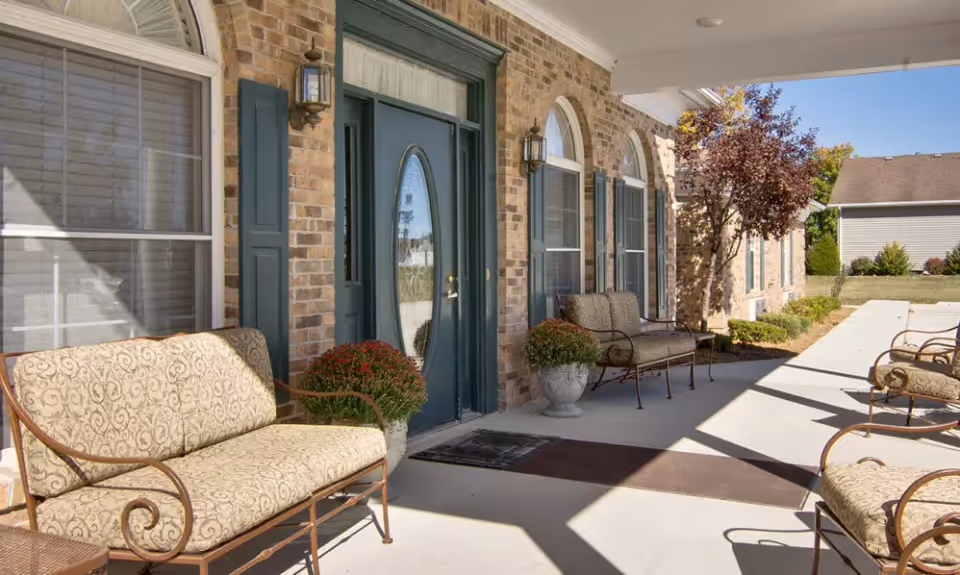 Outdoor covered patio area at Highland Crest Senior Living with cushioned metal benches, potted plants, brick exterior walls, green doors and windows with shutters, and a clear blue sky.