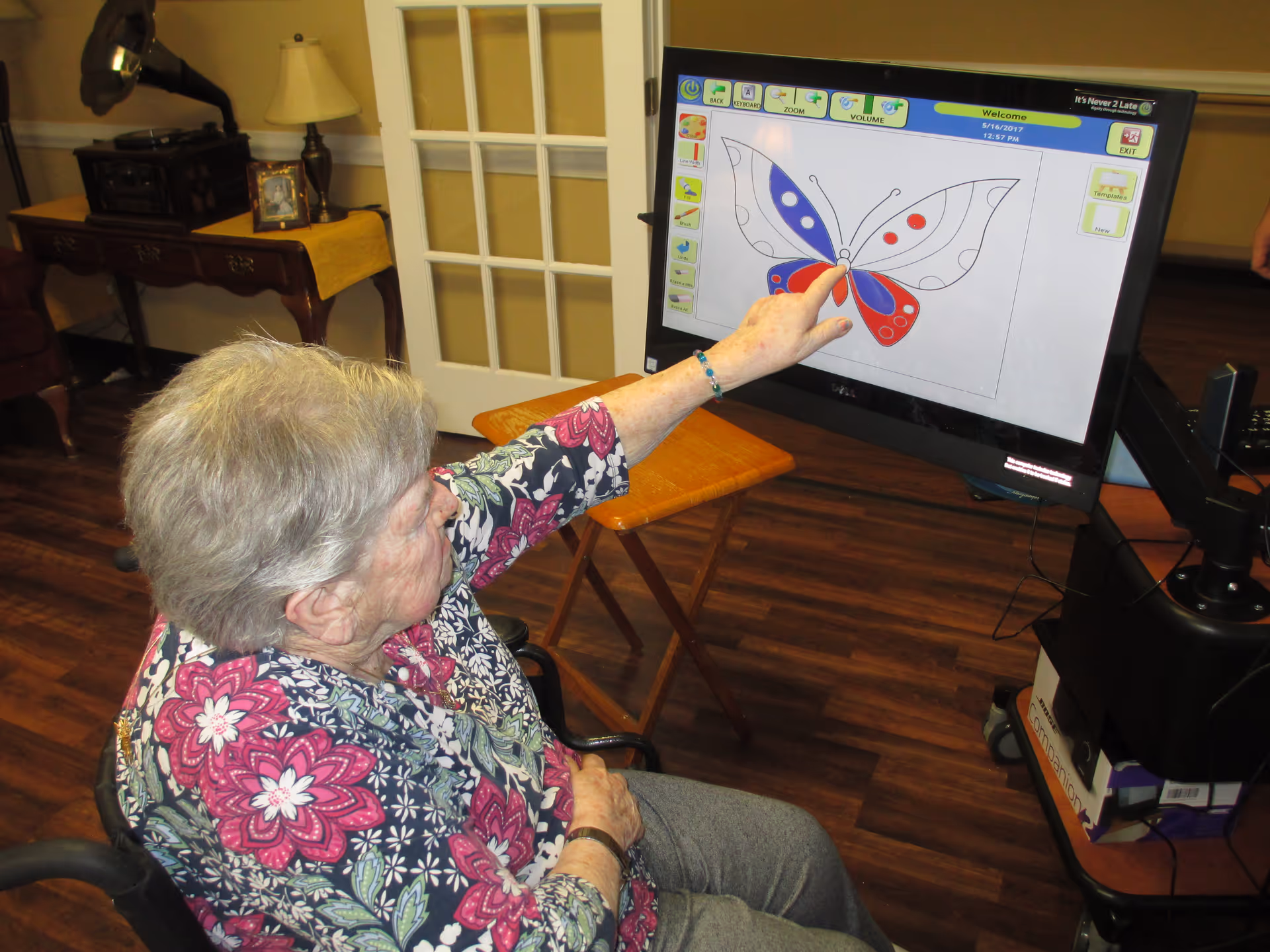 An elderly woman seated in a wheelchair is interacting with a touchscreen monitor displaying a butterfly coloring activity. The room has wooden flooring, a table with a lamp and a framed photo, and a door with glass panes in the background.