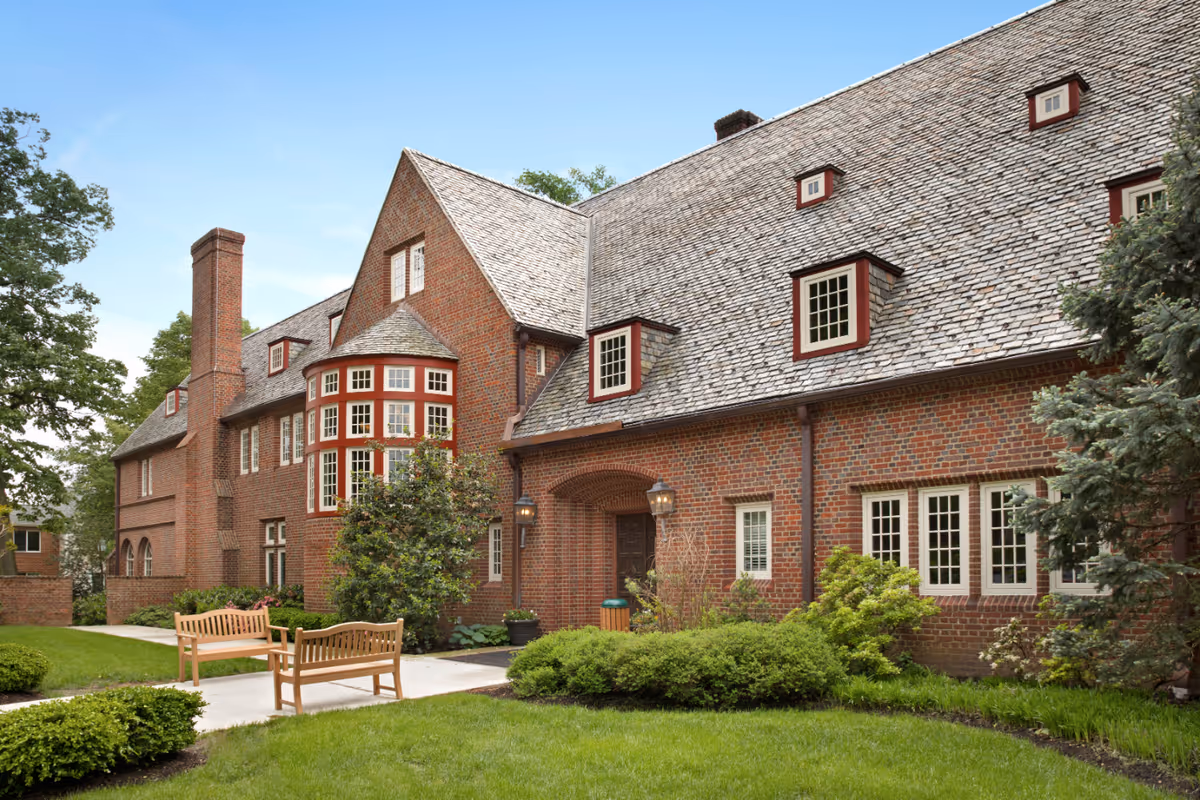 Exterior view of a large brick building with a steep shingled roof and multiple windows. The building is surrounded by well-maintained green lawns, shrubs, and trees. There are two wooden benches on a paved walkway in front of the building under a clear blue sky.