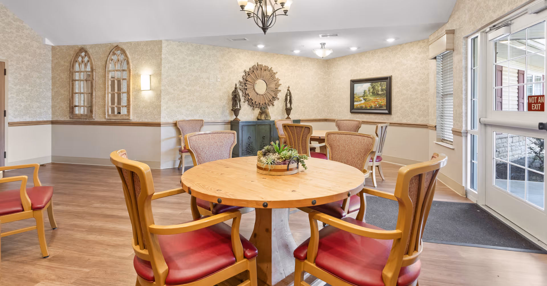 A bright and inviting common area in a senior living facility with a round wooden table surrounded by six wooden chairs with red cushions. The room has light-colored wallpaper, wooden flooring, and decorative elements including two arched mirrors on the left wall, a sunburst mirror above a small cabinet, two statues, and a framed landscape painting. There is a glass door on the right side with a 'Not An Exit' sign visible outside.