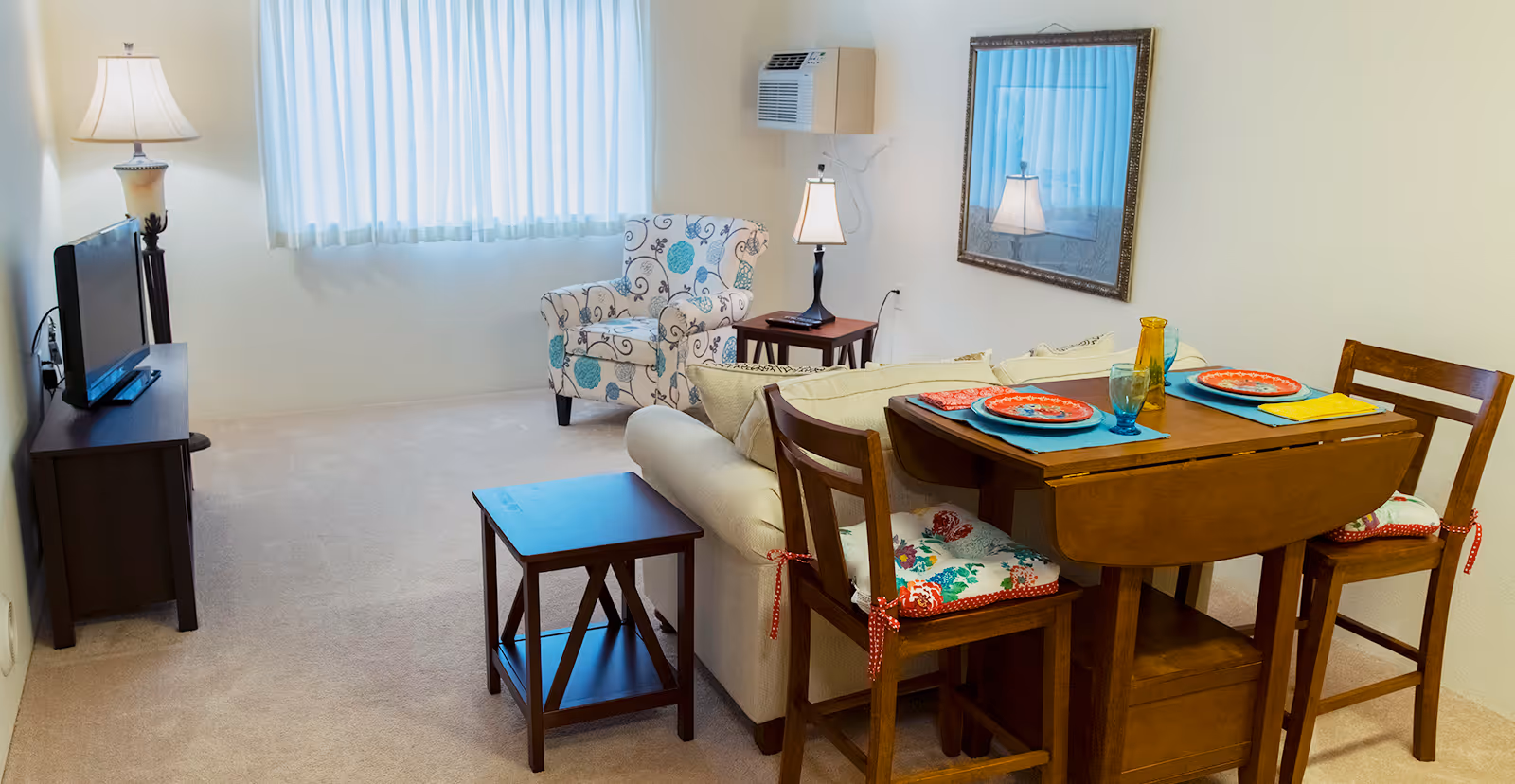 A cozy living room and dining area with a beige sofa, a floral patterned armchair, a wooden dining table set for two with colorful plates and glasses, a TV on a stand, a floor lamp, a small side table, and a wall-mounted air conditioner under a window with white curtains.