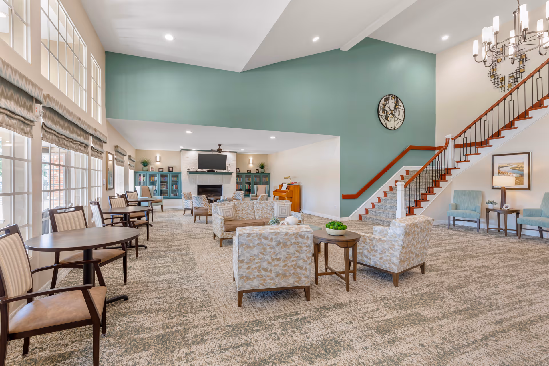 A spacious and well-lit living room area in a senior living facility with multiple seating arrangements including patterned armchairs, small round tables with chairs, and a sofa. The room features large windows with blinds, a fireplace with a TV mounted above it, built-in bookshelves, a piano, and a staircase with wooden handrails. The walls are painted in soft green and beige tones, and there is a modern chandelier hanging from the ceiling.