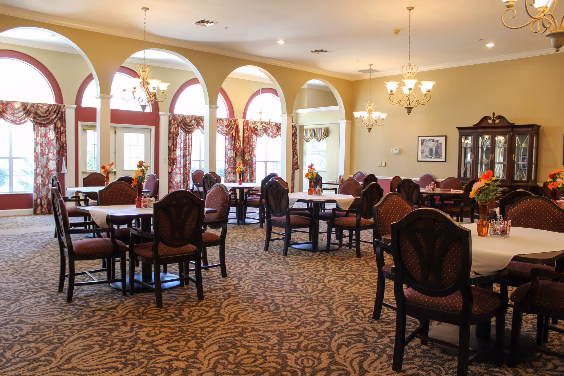 A spacious dining room with multiple round tables covered with white tablecloths, each surrounded by dark wooden chairs with red cushioned seats. The room features large arched windows with floral curtains, chandeliers hanging from the ceiling, and a patterned carpet. A wooden cabinet with glass doors is visible against one wall, and small flower arrangements are placed on each table.