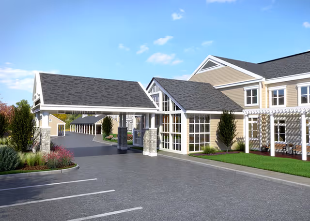 Exterior view of a senior living facility entrance with a covered drop-off area supported by stone pillars, adjacent to a building with large windows and beige siding. There is a parking lot in the foreground and landscaped greenery around the building under a clear blue sky.
