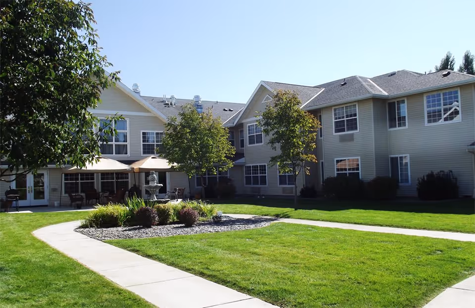 Exterior view of McKay Creek Assisted Living facility showing a two-story building with multiple windows, a well-maintained lawn, a curved concrete walkway, small trees, and a patio area with umbrellas and outdoor seating.