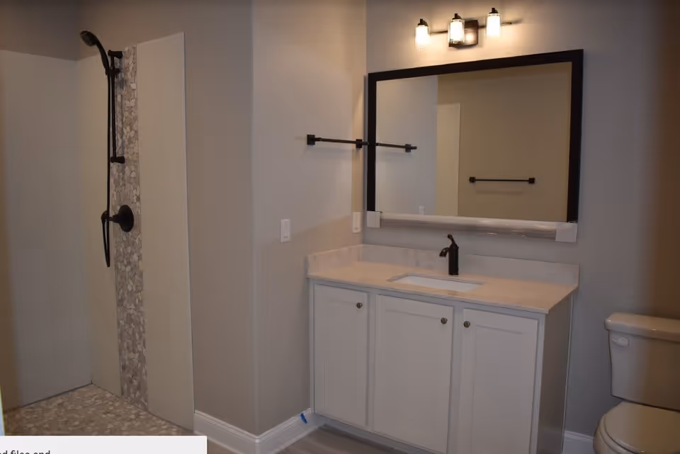 A modern bathroom featuring a walk-in shower with pebble tile flooring and a black showerhead, a white vanity with a marble countertop and an undermount sink, a large rectangular mirror with a black frame, a black faucet, and a toilet partially visible on the right side.