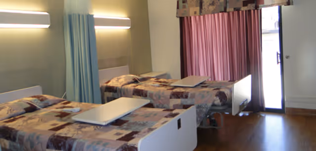 Two adjustable hospital-style beds with overbed tables in a shared patient room with curtains and wall lights.