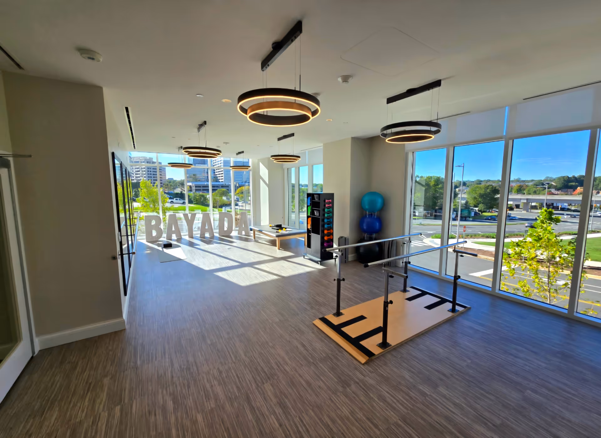 Bright and spacious rehabilitation or physical therapy room with large windows letting in natural light. The room features parallel bars for walking exercises, exercise balls stacked in a corner, a rack with colorful dumbbells, and a table with therapy equipment. Large letters spelling BAYADA are placed near the windows.