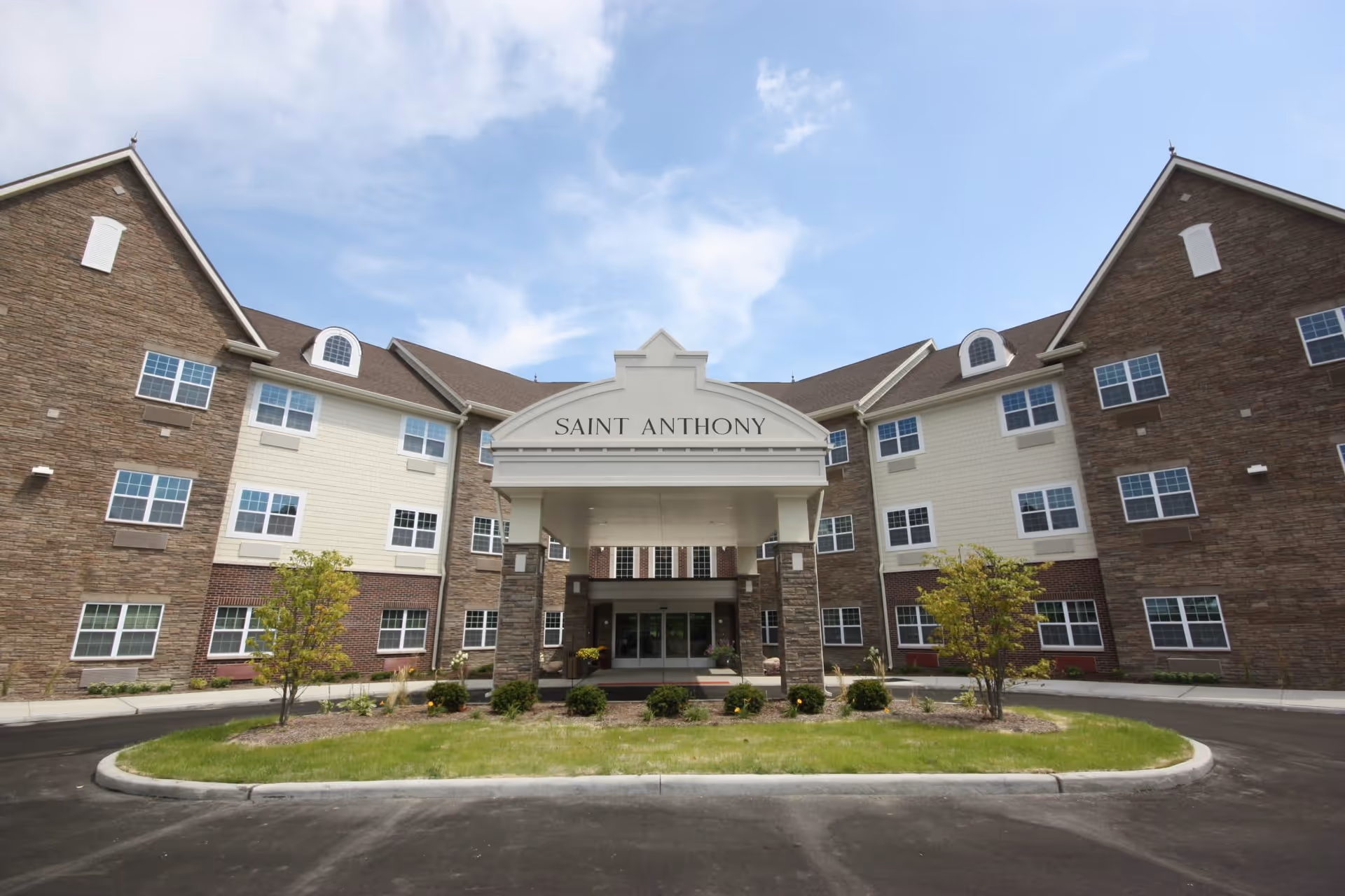 Front exterior view of a large senior living facility building with a covered entrance labeled 'Saint Anthony'. The building has multiple windows, stone and brick facade, and a circular driveway with landscaped greenery in front.