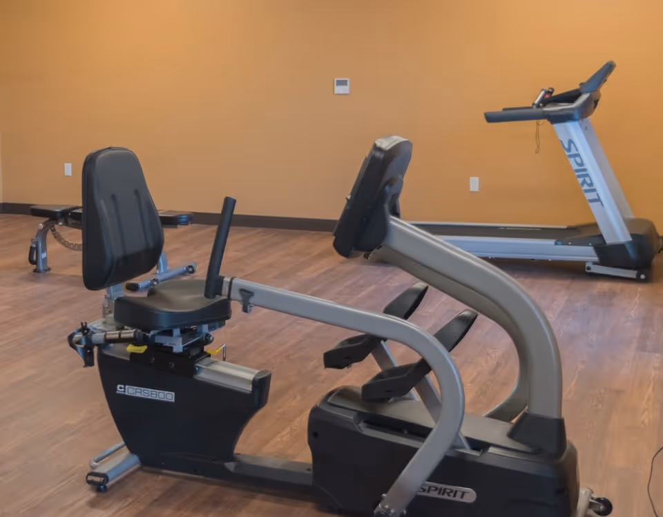 A small fitness room with a recumbent exercise bike in the foreground and a treadmill against a tan wall.