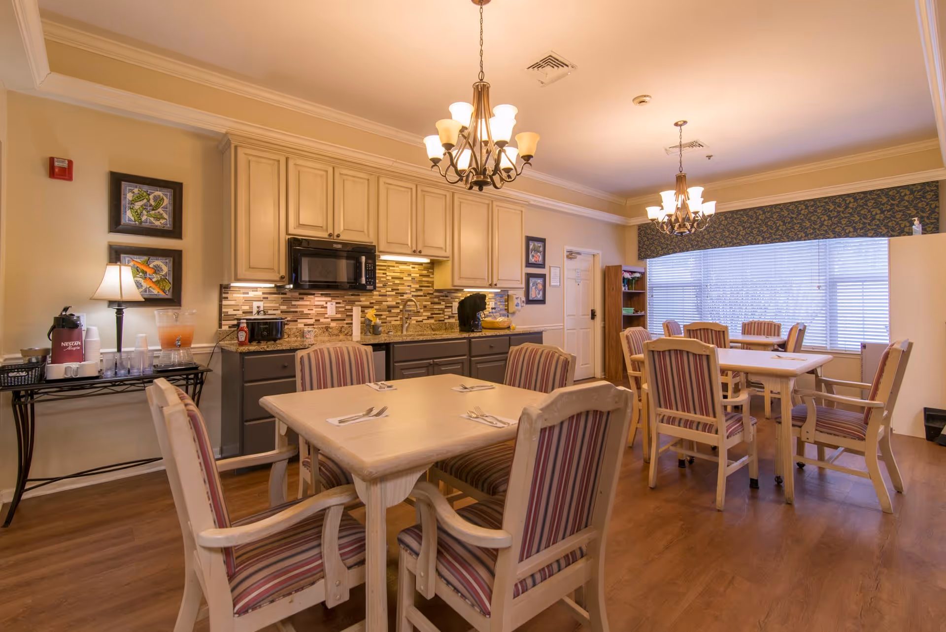 A dining room area in a senior living facility with several tables and chairs featuring striped upholstery. The room includes a kitchenette with cabinets, a microwave, a sink, and a coffee station. Two chandeliers hang from the ceiling, and a large window with blinds allows natural light to enter.