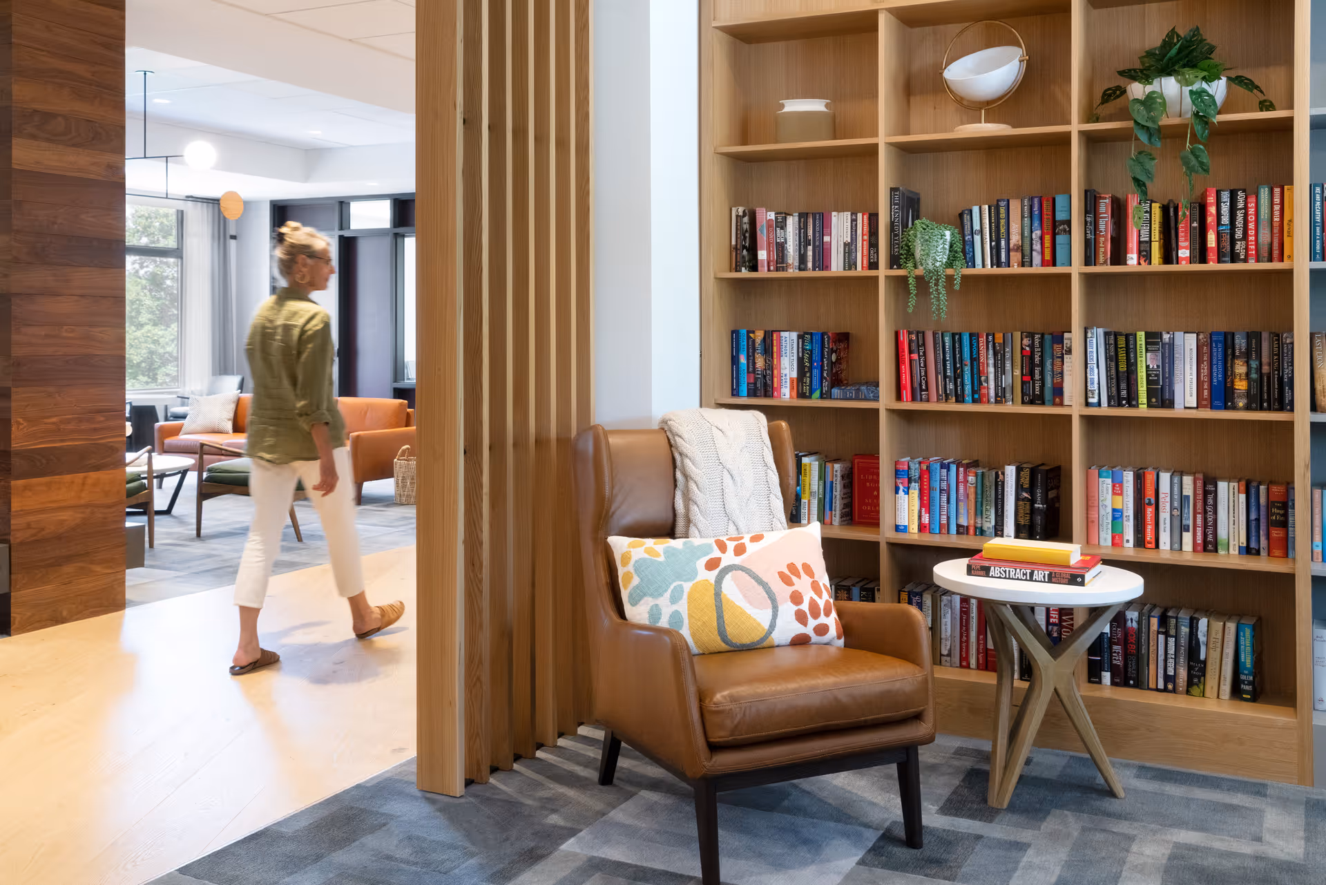 A cozy reading nook with a brown leather armchair adorned with a knitted throw and a colorful pillow, next to a small round table with books on it. Behind the chair is a wooden bookshelf filled with books and decorative plants. In the background, a woman walks through a modern, well-lit common area with large windows and comfortable seating.