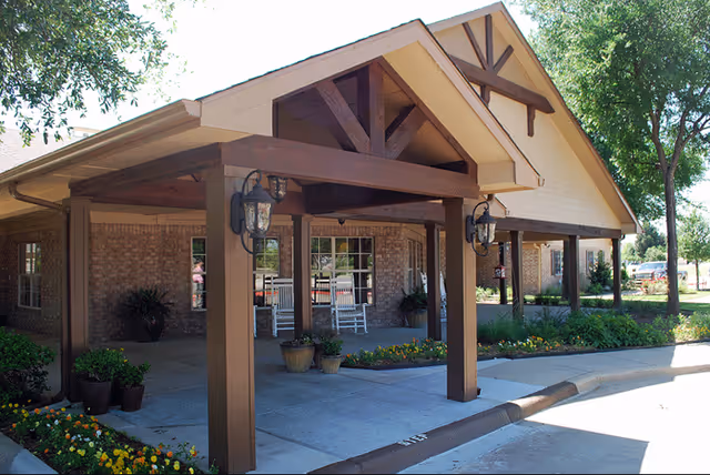 Covered porte-cochère with wooden beams and rocking chairs at the front entrance of a brick senior living building.