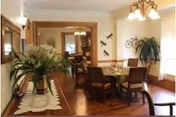 A warmly lit dining room with a wooden table and six chairs. The table is set with a centerpiece and placemats. There are plants and decorative wall hangings, including a bicycle sculpture on the wall. A sideboard with a floral arrangement and a mirror is visible on the left side. The room has hardwood floors and large windows with light curtains.