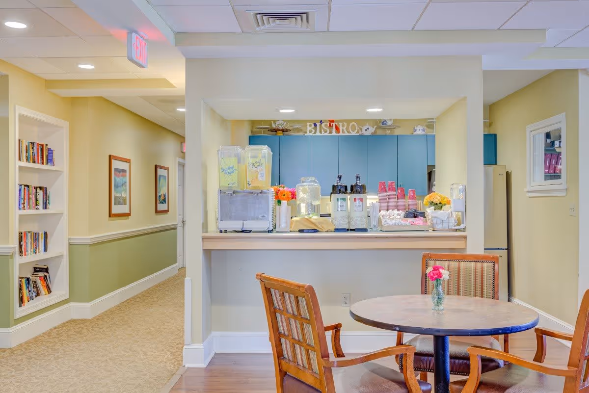 Interior view of a senior living facility featuring a small bistro counter with beverage dispensers, cups, and flowers. In front of the counter is a round table with four wooden chairs, one with a small vase holding a pink flower. To the left, there is a hallway with framed pictures on the wall and built-in bookshelves filled with books. The walls are painted in light yellow and green tones.