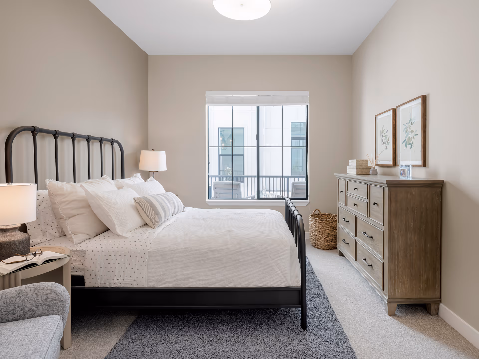A neatly made bed with white bedding and multiple pillows in a bedroom with beige walls. There is a black metal bed frame, a wooden dresser with decorative items and framed artwork above it, a woven basket on the floor, and a window showing an exterior view. A bedside table with a lamp and an open book is visible on the left side.