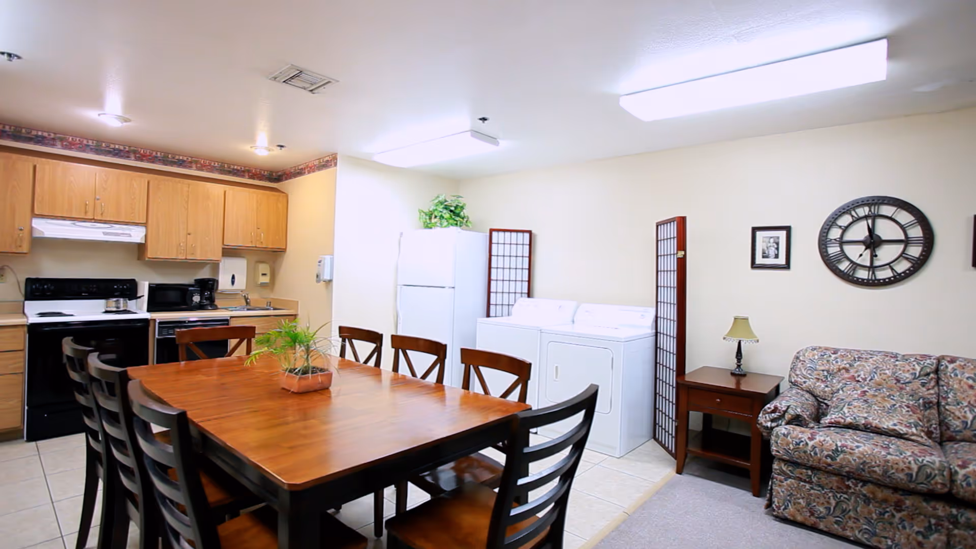 A room featuring a wooden dining table with eight chairs, a kitchen area with wooden cabinets, a stove, microwave, and sink, a white refrigerator, a washer and dryer, a small side table with a lamp, a floral-patterned couch, a large wall clock, and framed pictures on the wall.