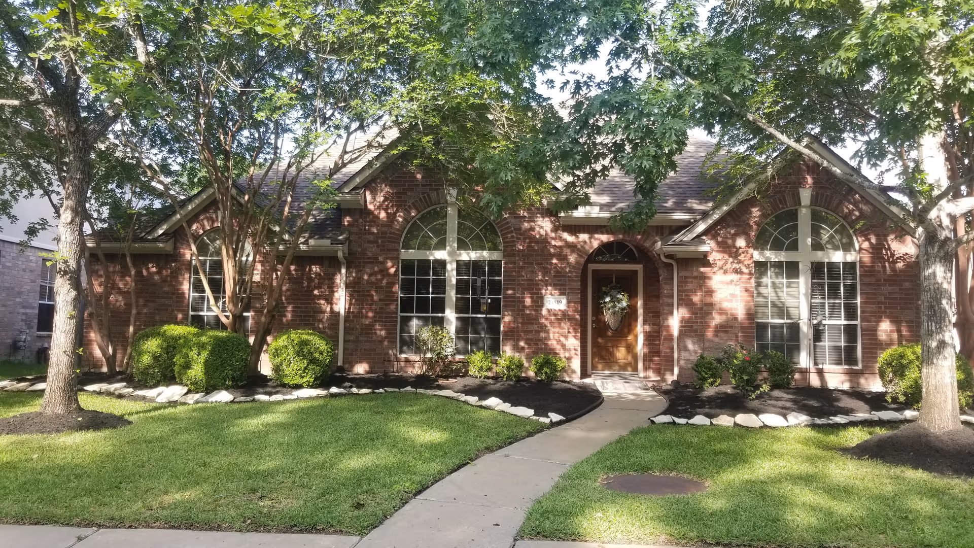 Front exterior view of a single-story brick house with a wooden front door decorated with a wreath. The house has large arched windows and is surrounded by green grass, bushes, and trees casting shadows on the facade. A concrete pathway leads to the front door.