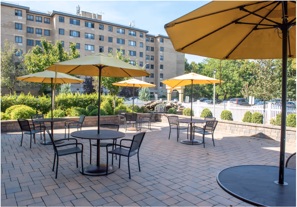 Outdoor patio area with several round tables and black metal chairs, each table shaded by a large yellow umbrella. The patio is paved with bricks and surrounded by greenery, with a multi-story building and parked cars visible in the background under a clear blue sky.