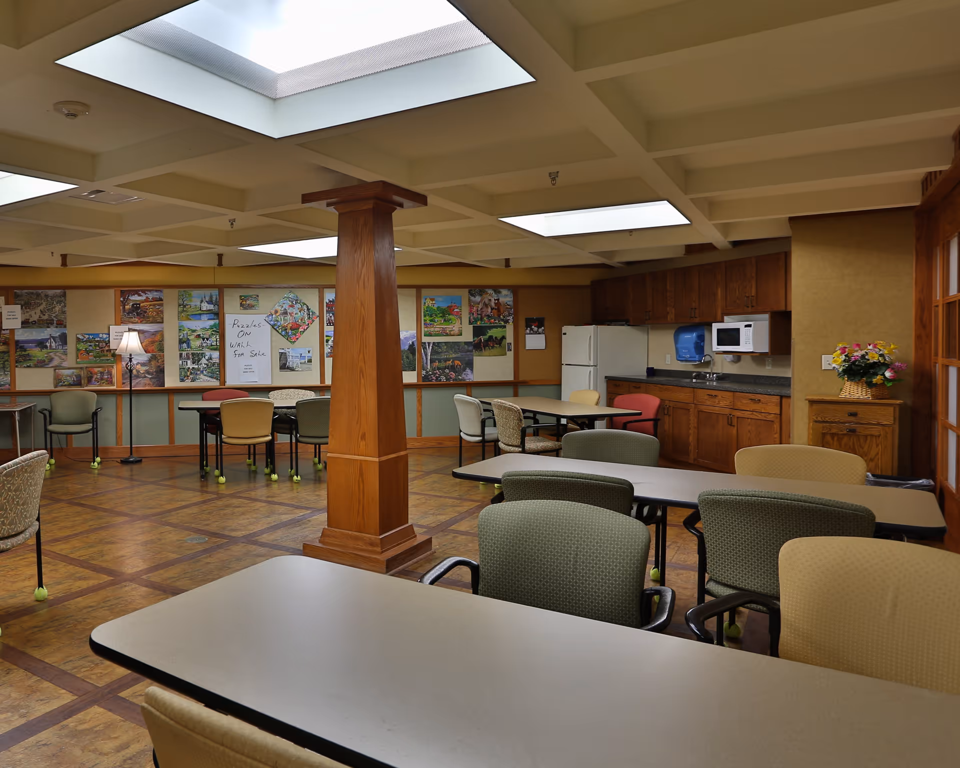 A well-lit common room with tables and chairs arranged for group activities or dining. The room features a wooden column in the center, a kitchenette area with a refrigerator, microwave, and sink, and a bulletin board with various pictures and a sign that reads 'Puzzles ON WALL For Sale'. The ceiling has large recessed lighting panels, and there is a floor lamp in the corner.