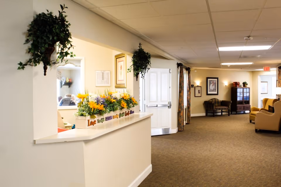 Interior view of a senior living facility hallway with beige walls and carpet. There is a counter with colorful artificial flowers and decorative plants hanging above it. The hallway has framed pictures on the walls, a wooden cabinet, and several upholstered chairs arranged for seating. An exit sign is visible in the background.