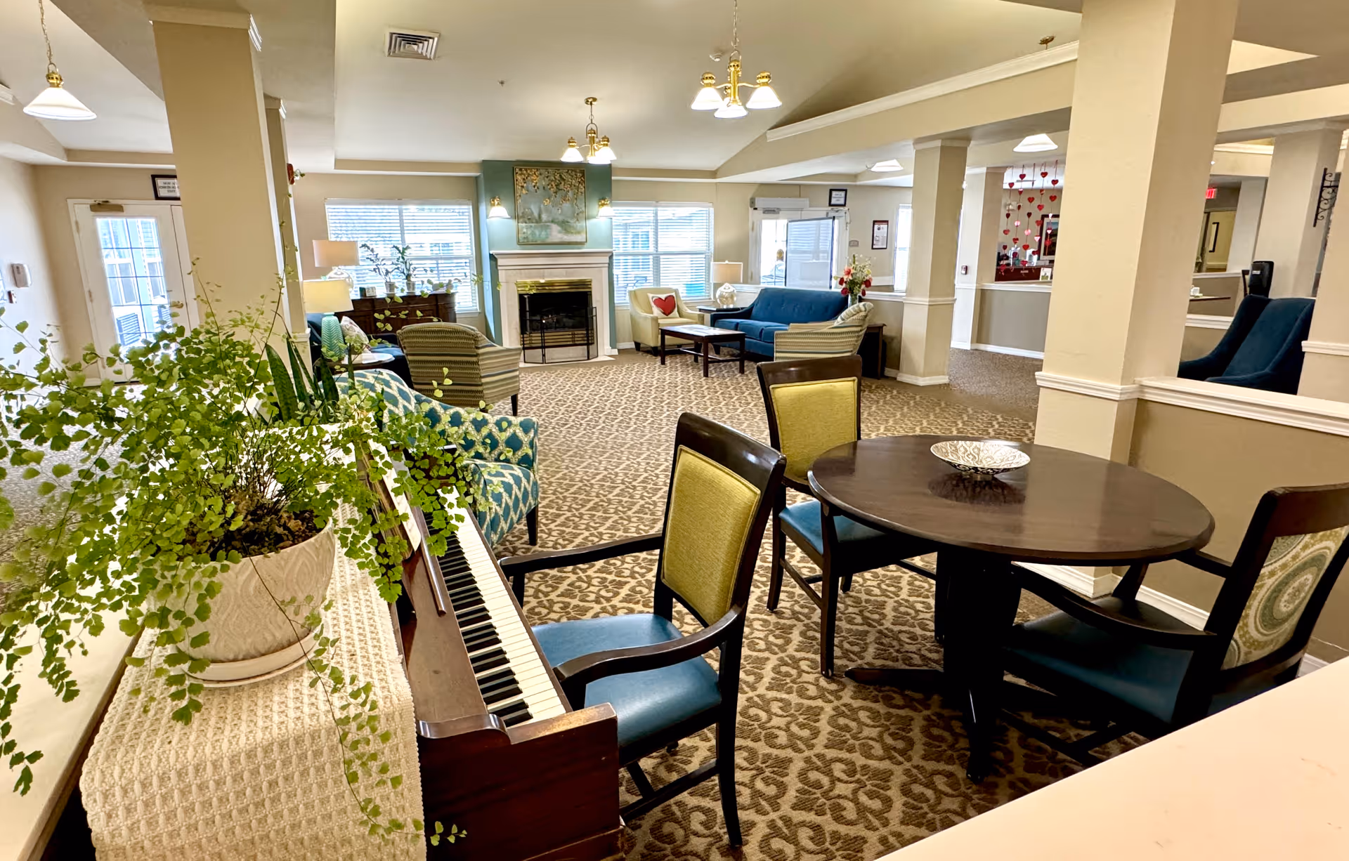 A cozy senior living facility common area with patterned carpet, a piano with a potted plant on top, a round wooden table with four chairs, and a seating area with armchairs and a sofa near a fireplace. The room is well-lit with natural light from large windows and ceiling lights.