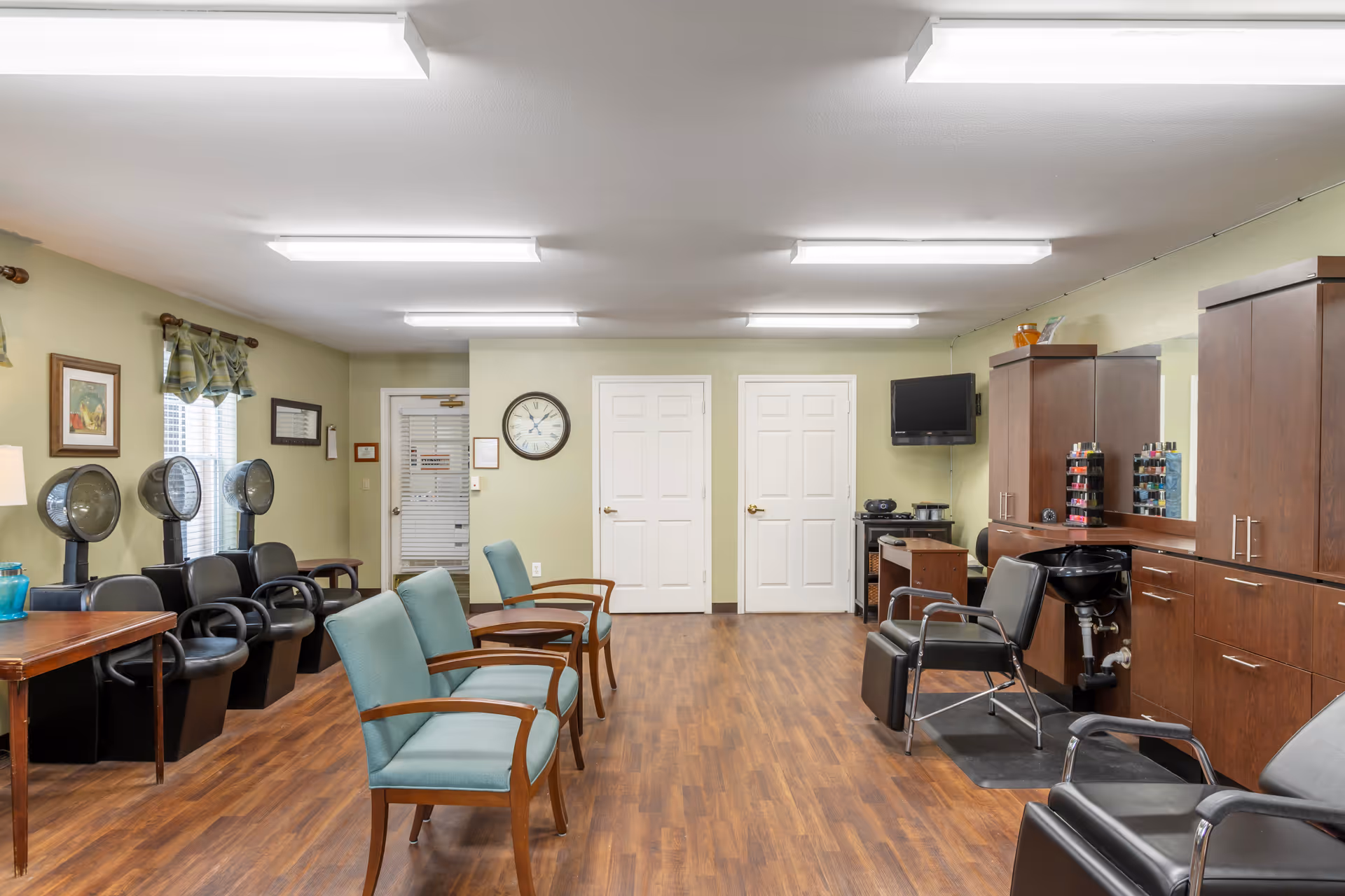 Interior of a salon room in a senior living facility with multiple black salon chairs and hair dryers on the left, teal waiting chairs in the center, and a hair washing station with a black chair and sink on the right. The room has wood flooring, light green walls, a wall clock, a mounted TV, and wooden cabinets with nail polish displays.