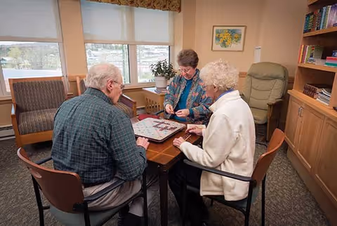 Three elderly people sitting around a wooden table playing a board game in a well-lit room with large windows, bookshelves, and comfortable chairs.