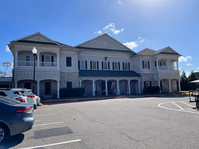 Front exterior view of a two-story building with a brick and light-colored facade, featuring multiple windows with shutters, balconies, and a covered entrance. Several cars are parked in the parking lot in front of the building under a clear blue sky.