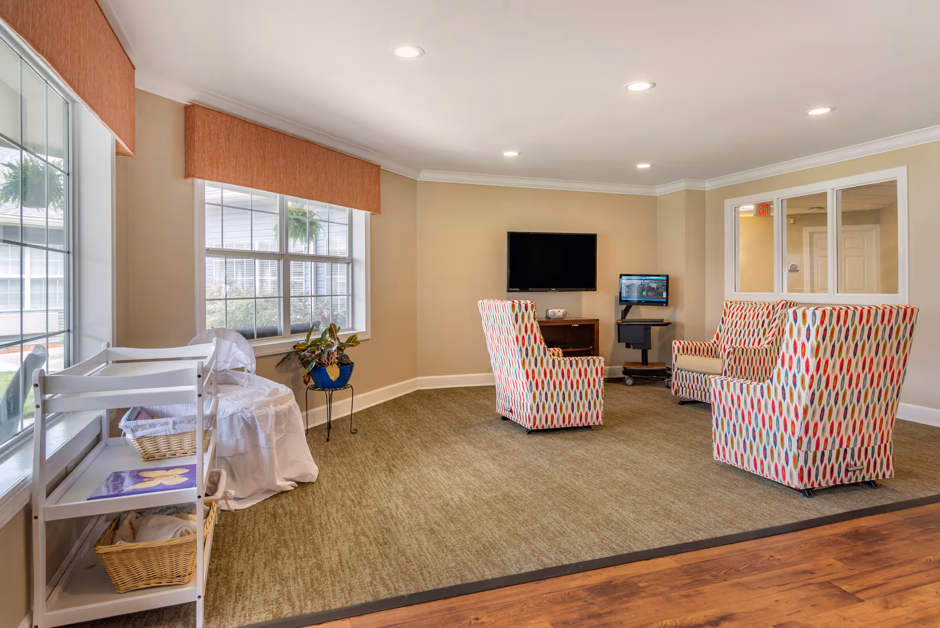 A cozy sitting area in a senior living facility with three colorful patterned armchairs arranged around a wall-mounted flat screen TV and a smaller monitor on a stand. The room has beige walls, a large window with an orange valance, a small table with a potted plant, and a white shelving unit with baskets and linens. The floor is carpeted with a greenish tone, and there is wood flooring in the foreground.