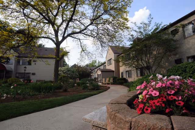 A paved walkway curves through a landscaped garden area with green grass, trees, and colorful flowers in a raised stone planter. Beige two-story buildings with windows and brown roofs are visible on both sides of the path under a partly cloudy sky.