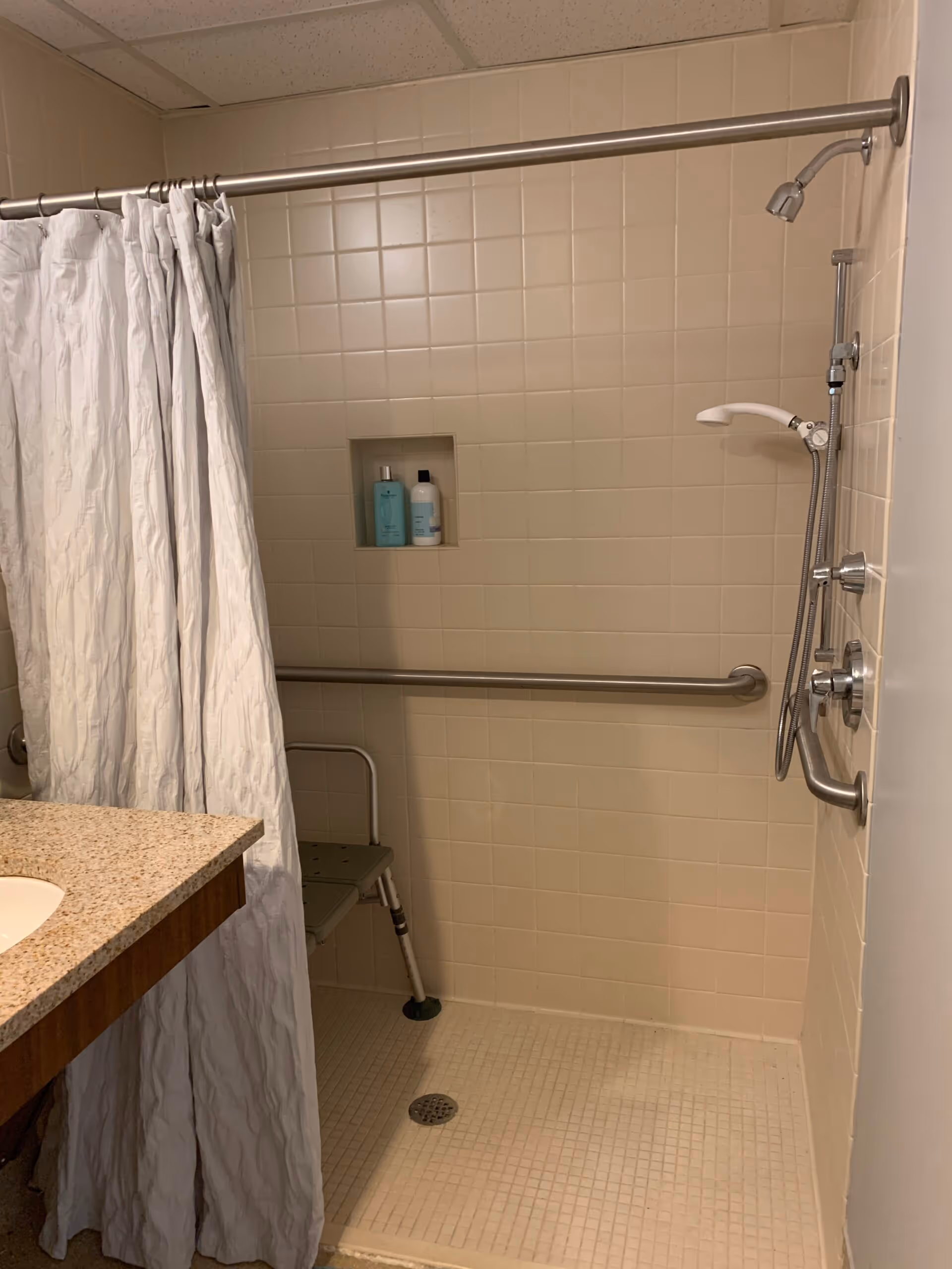 A beige tiled shower area with a white shower curtain partially drawn to the left. Inside the shower, there is a metal grab bar along the back wall, a handheld showerhead attached to a vertical bar, and a small shower chair. Two bottles of shower products are placed in a recessed shelf in the wall. To the left of the shower is a countertop with a sink.