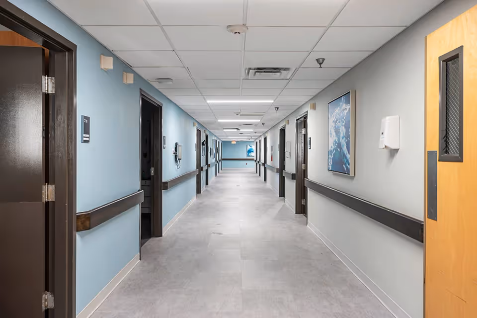 Long institutional hallway with blue-gray walls, handrails, multiple room doors, ceiling tiles and wall art.