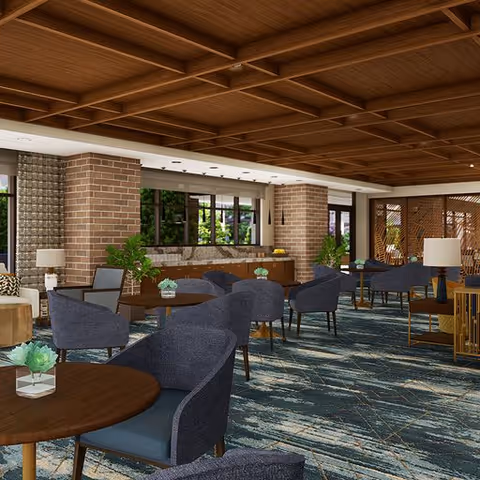 A spacious and well-lit lounge area with multiple round wooden tables surrounded by dark upholstered chairs. The room features a coffered wooden ceiling, brick columns, large windows with views of greenery outside, and decorative plants placed around. The carpet has a blue and beige abstract pattern, and there are modern table lamps on side tables.