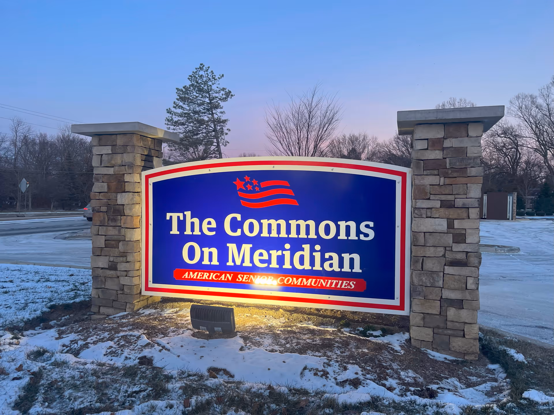 Illuminated entrance sign reading "The Commons On Meridian" set between two stone pillars with snow on the ground at dusk.