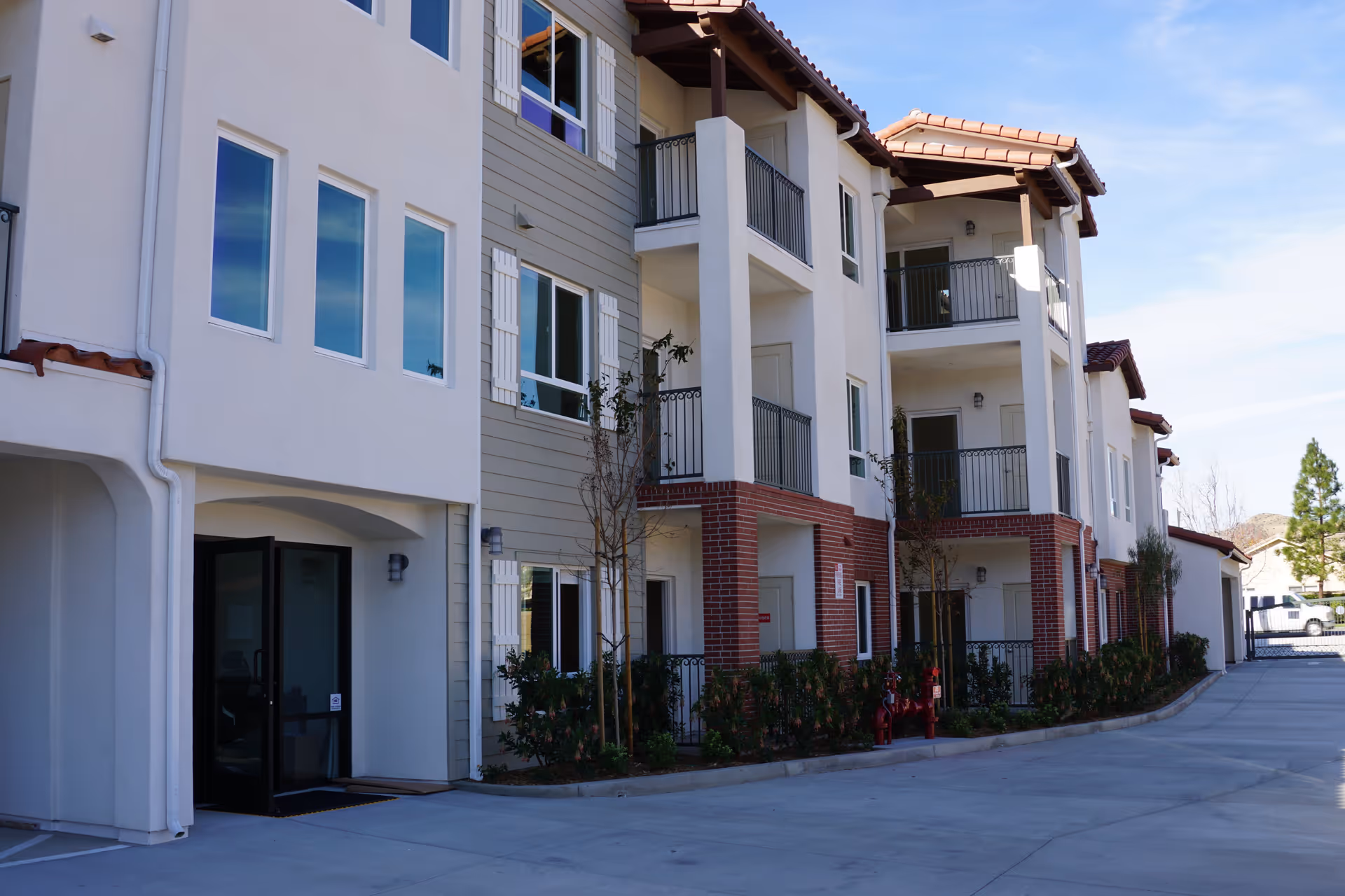 Exterior view of a multi-story residential building with balconies, windows, and a driveway. The building features a combination of white and beige walls with red brick accents and a tiled roof. Small trees and shrubs are planted along the building's base.