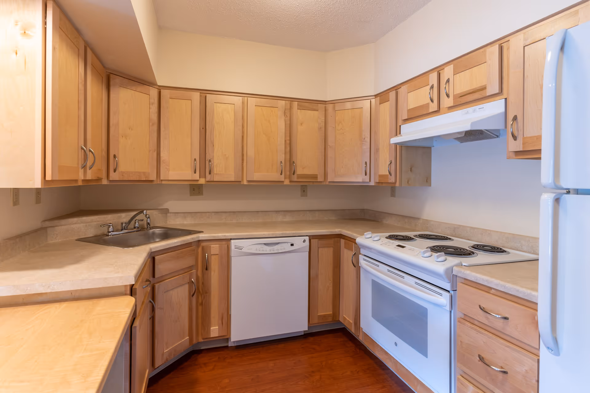 A kitchen with light wood cabinets, beige countertops, a stainless steel sink, a white electric stove with four coil burners, a white range hood, a white dishwasher, and a white refrigerator. The floor is a medium-tone wood.