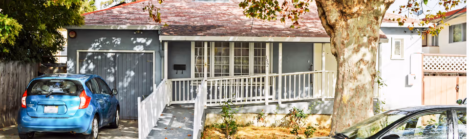 Front exterior view of a single-story residential building with a gray facade, a sloped roof, a wheelchair accessible ramp leading to the front door, and two cars parked in the driveway. There is a large tree in the front yard.