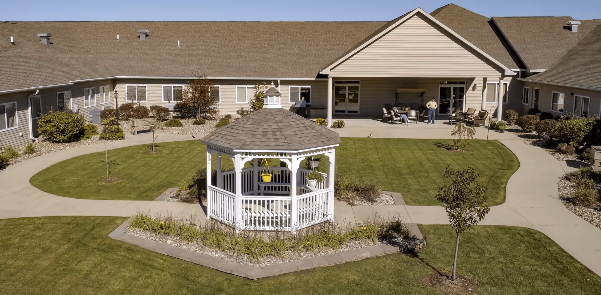 White gazebo in the center of a grassy courtyard with walkways and a one-story assisted living building in the background.