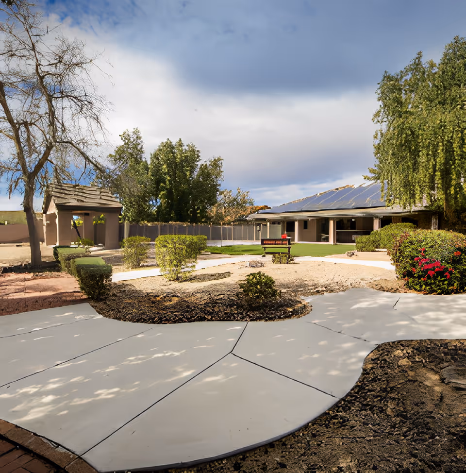 Outdoor garden area at Sweetwater Groves By Majestic Residences featuring a paved walkway, trimmed bushes, a bench, trees, and a building with a covered patio in the background under a partly cloudy sky.