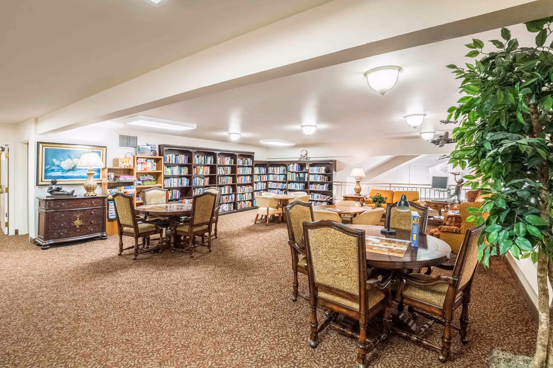 A spacious and well-lit library and lounge area in a senior living facility featuring multiple bookshelves filled with books, several round tables with upholstered chairs, a large potted plant, and comfortable seating with lamps and artwork on the walls.