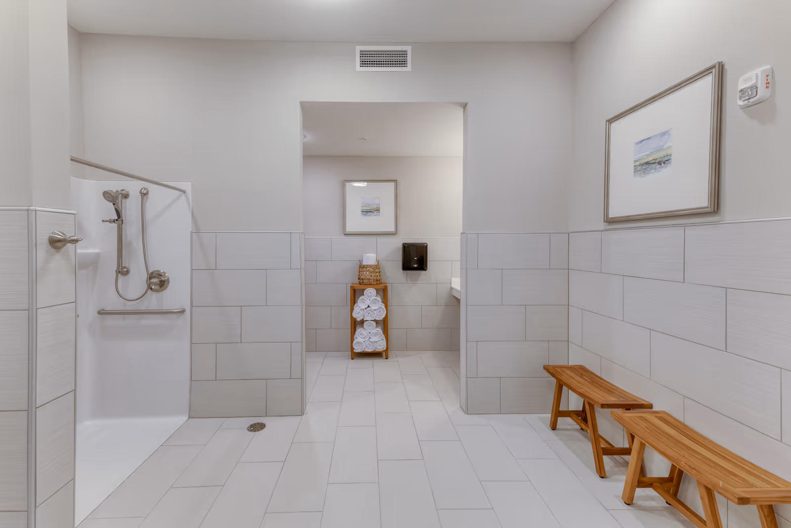 A clean, modern shower room with light gray tiled walls and floor. The shower area on the left has a handheld showerhead and grab bars. In the center background, there is a wooden rack holding neatly rolled white towels and a basket on top. On the right side, two wooden benches are placed against the wall, which is decorated with a framed picture. The room is well-lit with a minimalist design.