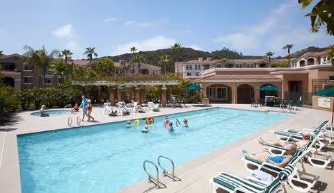 Community outdoor swimming pool with lounge chairs, people swimming and sunbathing, and surrounding Mediterranean-style buildings and palm trees.